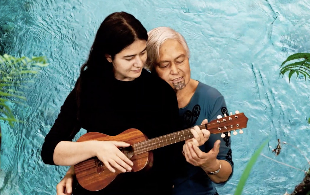 A girl sits on the knee of an older wāhine, playing the guitar together. As they sit on a chair together playing, the background shows a river flowing with blue water.